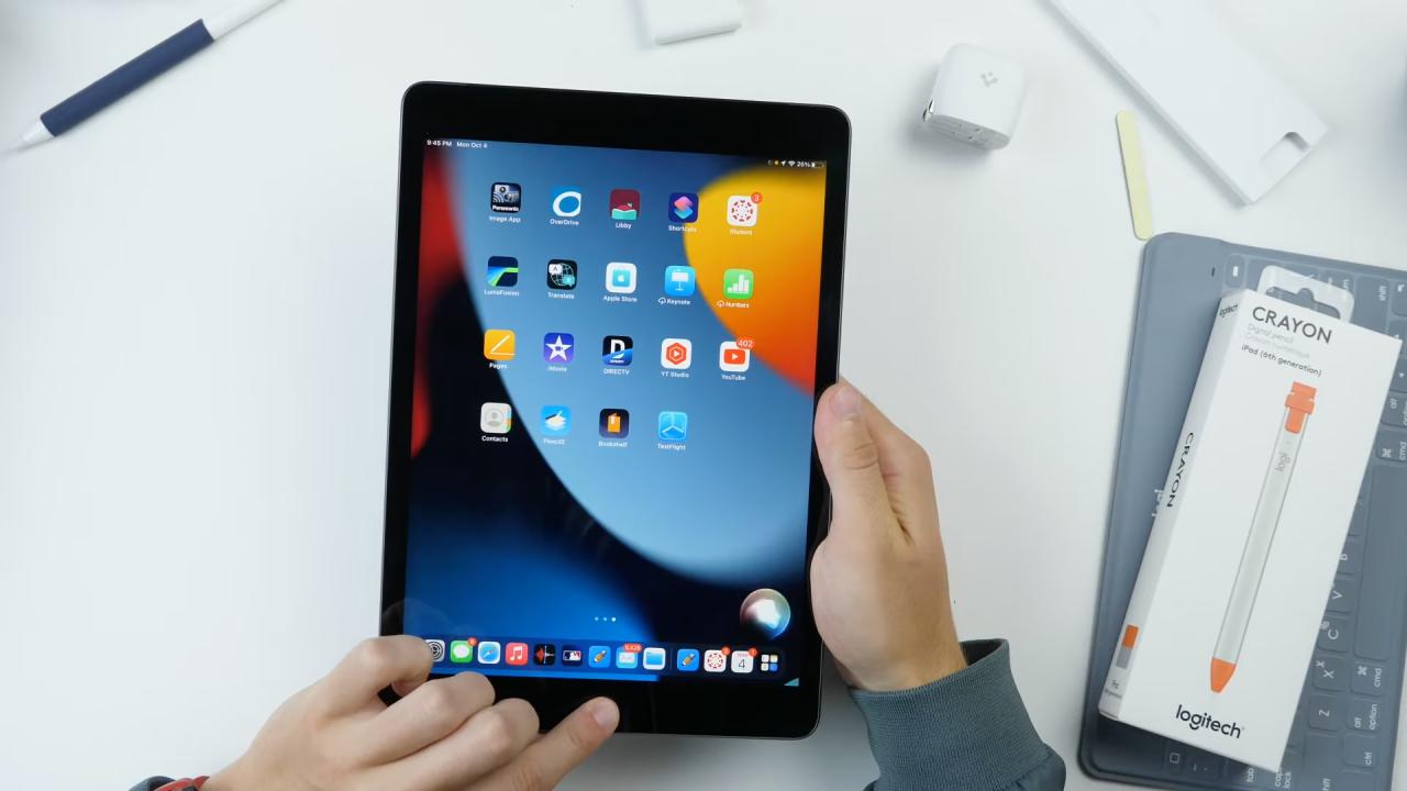 Una vista de arriba hacia abajo de un escritorio de trabajo blanco con las manos de un hombre sosteniendo el iPad de novena generación de Apple en las manos con la pantalla de inicio predeterminada que se muestra en la pantalla