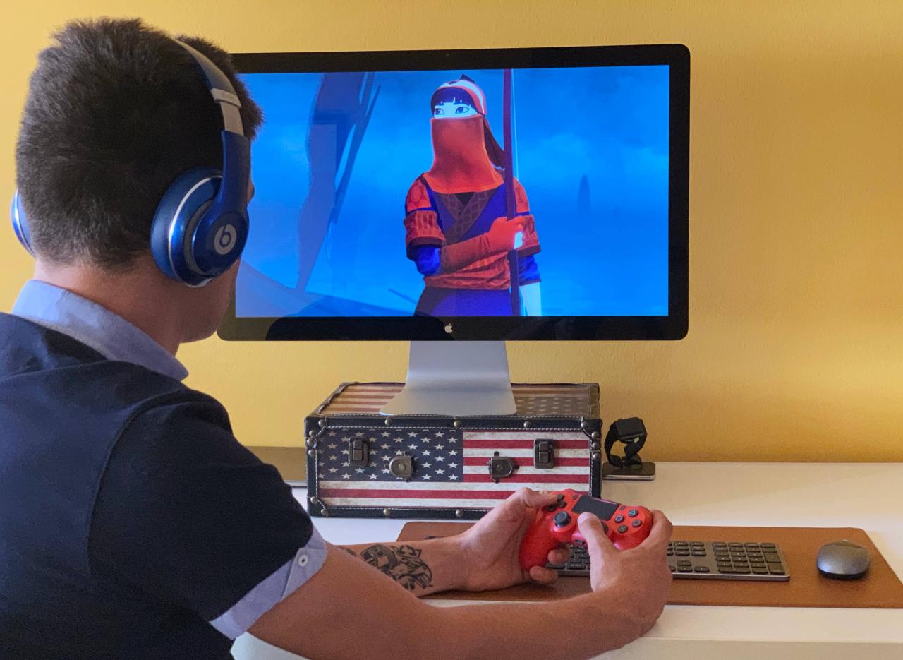 Una fotografía que muestra a un hombre joven sentado en su escritorio, sosteniendo un controlador PlayStation DualShock rojo en la mano y jugando un juego de Apple Arcade en su Apple Thunderbolt Display
