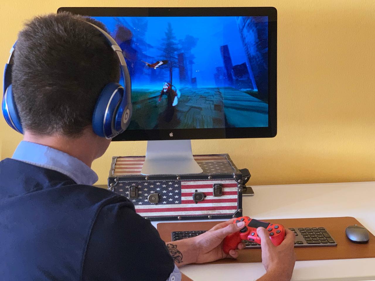 Una foto que muestra a un hombre joven de atrás que se sienta en un escritorio y sostiene un control en sus manos, jugando un juego en su Mac