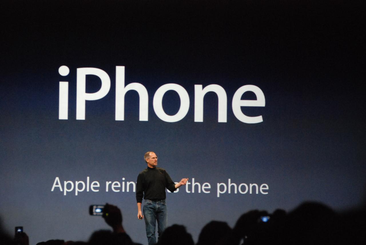 Steve Jobs de pie frente a una diapositiva en la presentación del iPhone de enero de 2007 que muestra el lema "Apple reinventa el teléfono"